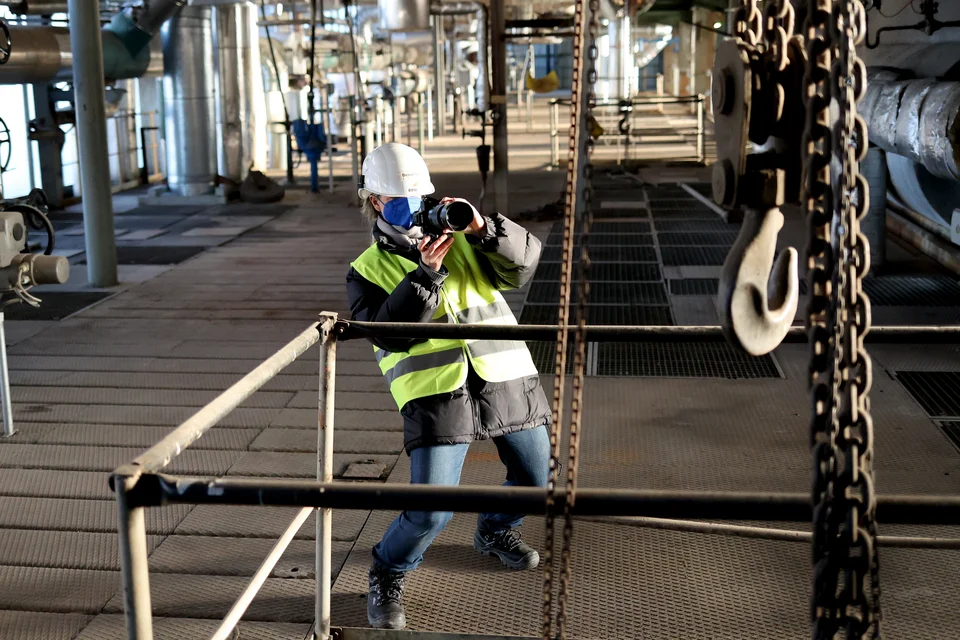 Fotografin Jessica Backhaus bei der Arbeit in einer Industrieanlage. Sie trägt Helm, Warnweste und Maske und fotografiert durch die Gitterstruktur einer Empore. Im Vordergrund sind schwere Ketten und ein großer Metallhaken sichtbar.