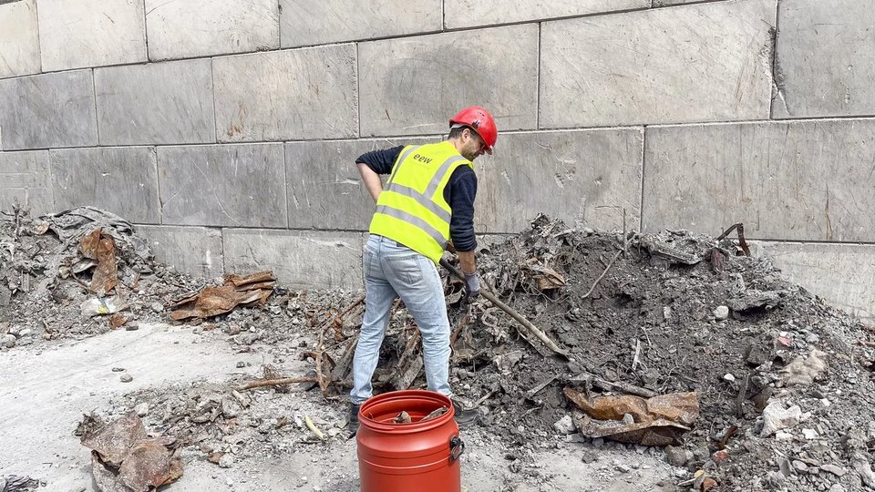 Fotograf Sjoerd Knibbeler bei der Arbeit am EEW-Standort in Delfzijl (Niederlande). Er trägt Sicherheitsweste und Helm und durchsucht mit einem Werkzeug einen Haufen aus Schlacke und Metallresten.