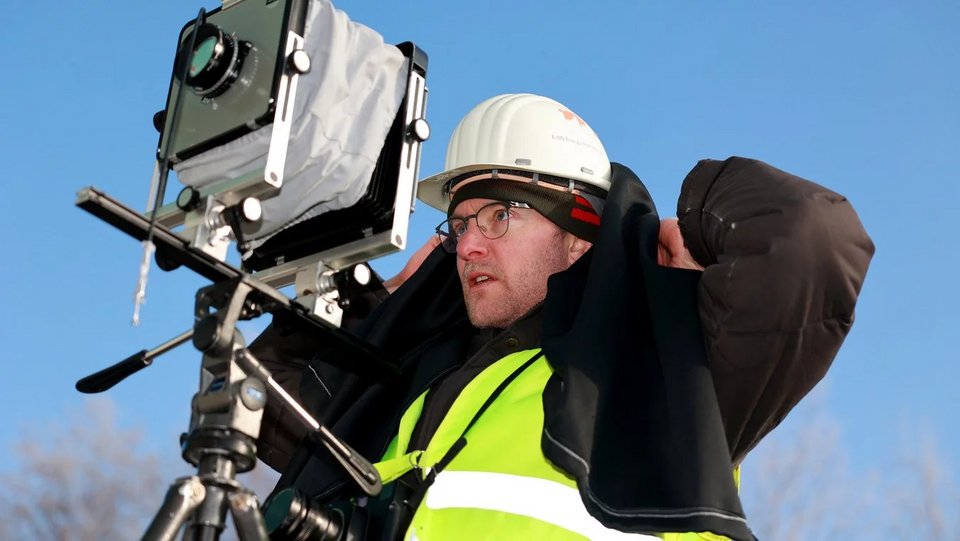 Fotograf Lukas Hoffmann bei der Arbeit mit einer Großbildkamera auf Stativ. Er trägt Helm und Warnweste und bereitet sich darauf vor, unter dem schwarzen Tuch die Kamera zu justieren.
