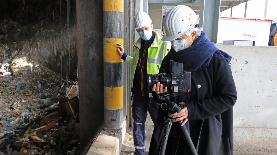 Fotografin Friederike von Rauch bei der Arbeit mit einer Mittelformatkamera auf Stativ in einer Abfallverwertungsanlage. Sie trägt Helm und Maske, neben ihr steht eine Person in Warnweste vor einem großen Müllbunker.